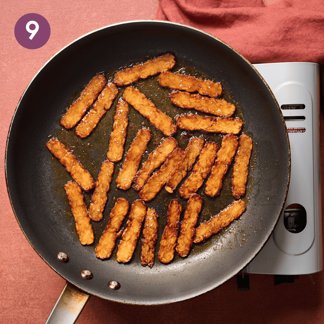 Darker tempeh bacon slices cooking on the stovetop.