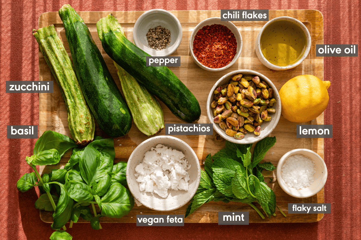 ingredients for zucchini salad laid out on wooden cutting board on red background. 