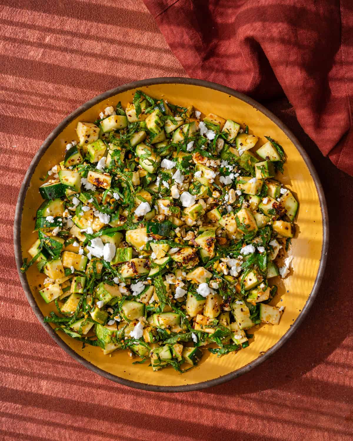 zucchini salad tossed with chili oil, pistachios, feta, basil, and mint in a yellow bowl on a red surface.