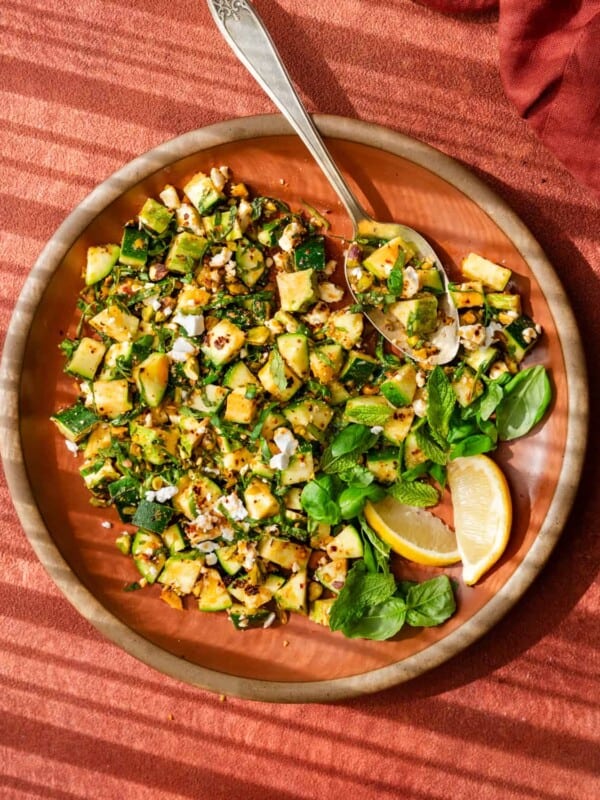 zucchini salad tossed with chili oil, pistachios, feta, basil, and mint on a red place on a red surface.