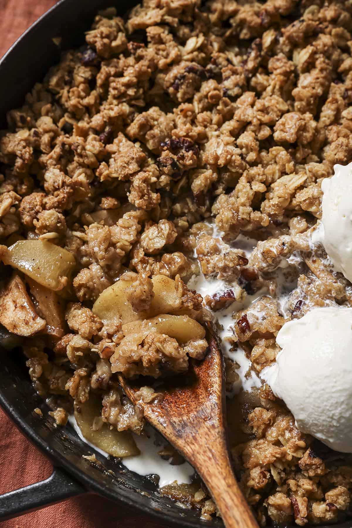 Apple crisp in a cast-iron pan being scooped out with a wooden spoon beside melting vanilla ice cream.