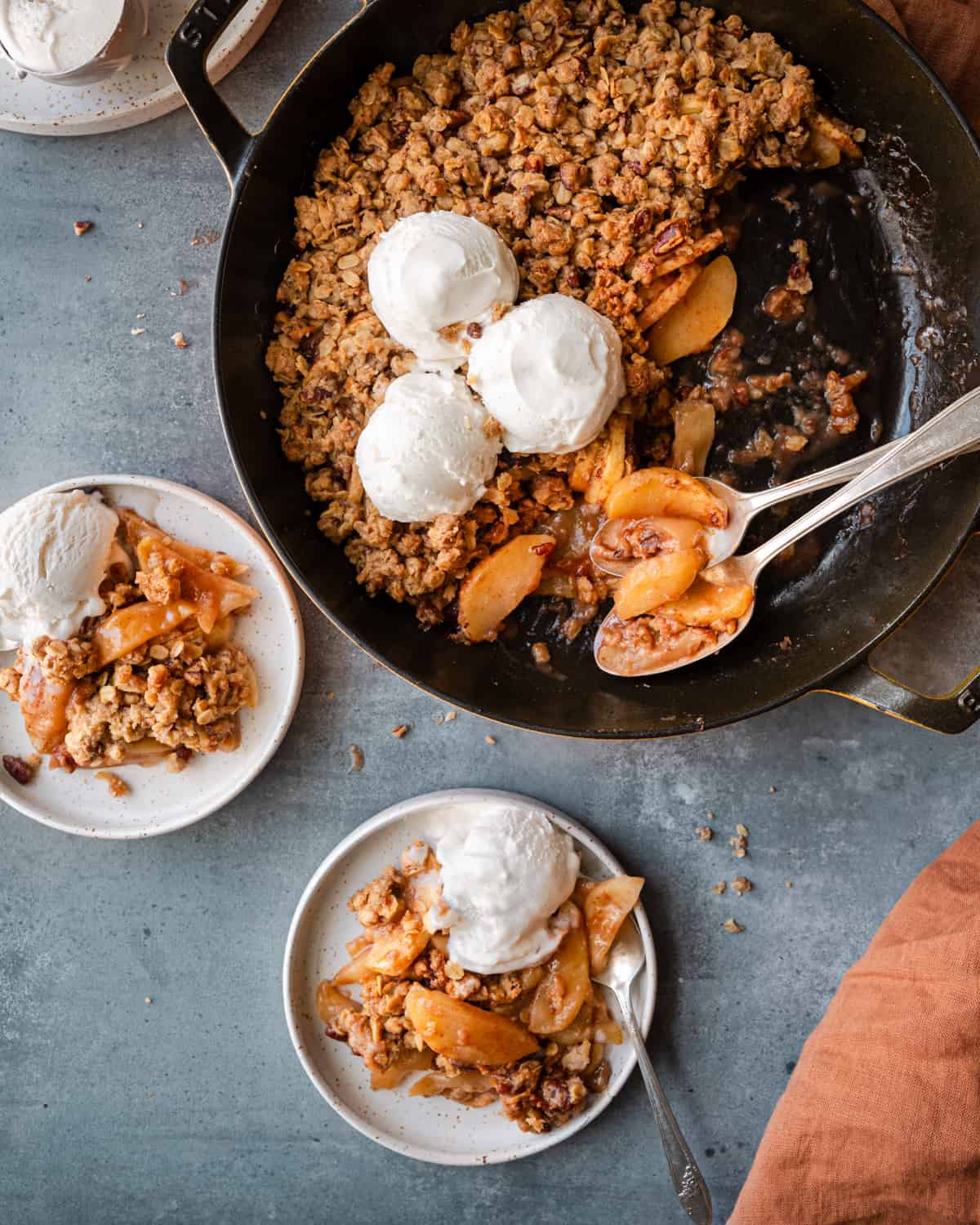 Two plates of apple crisp with ice cream next to skillet of apple crisp on a table.