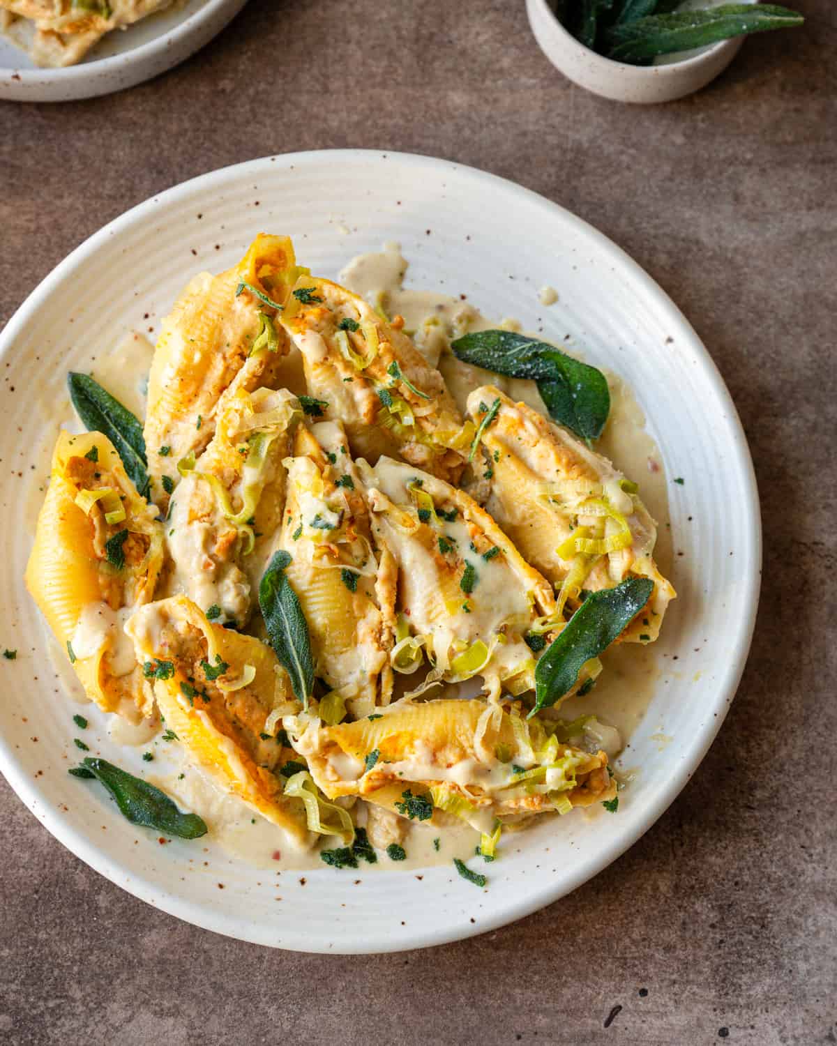 pumpkin ricotta stuffed jumbo shells on a speckled white plate, garnished with cream sauce and fried sage.