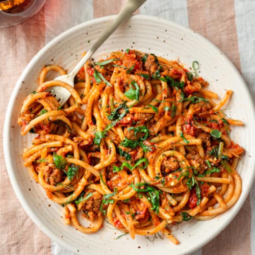 bucatini pasta with sausage tomato sauce, garnished with basil on a speckled plate on a striped napkin.