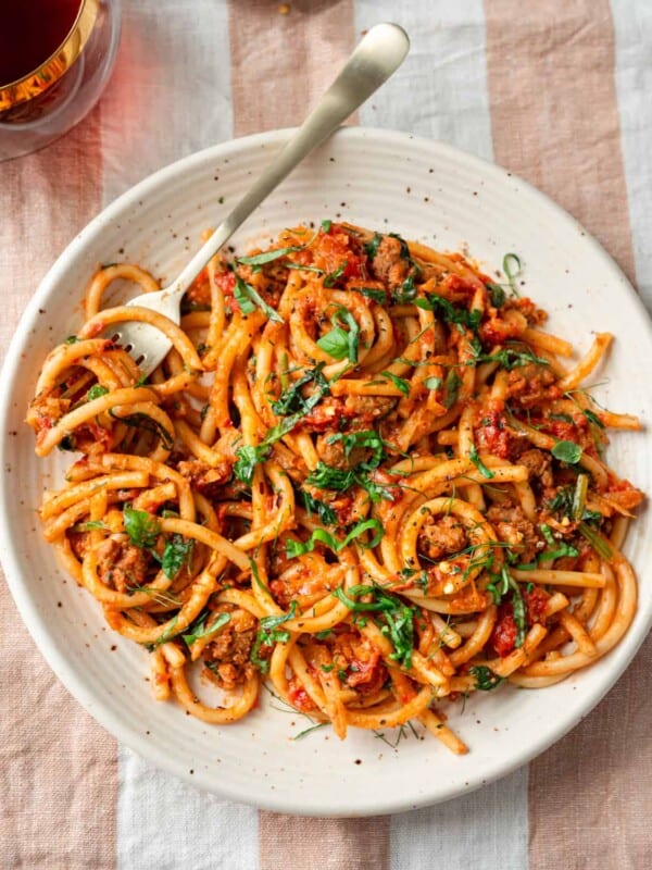 bucatini pasta with sausage tomato sauce, garnished with basil on a speckled plate on a striped napkin.