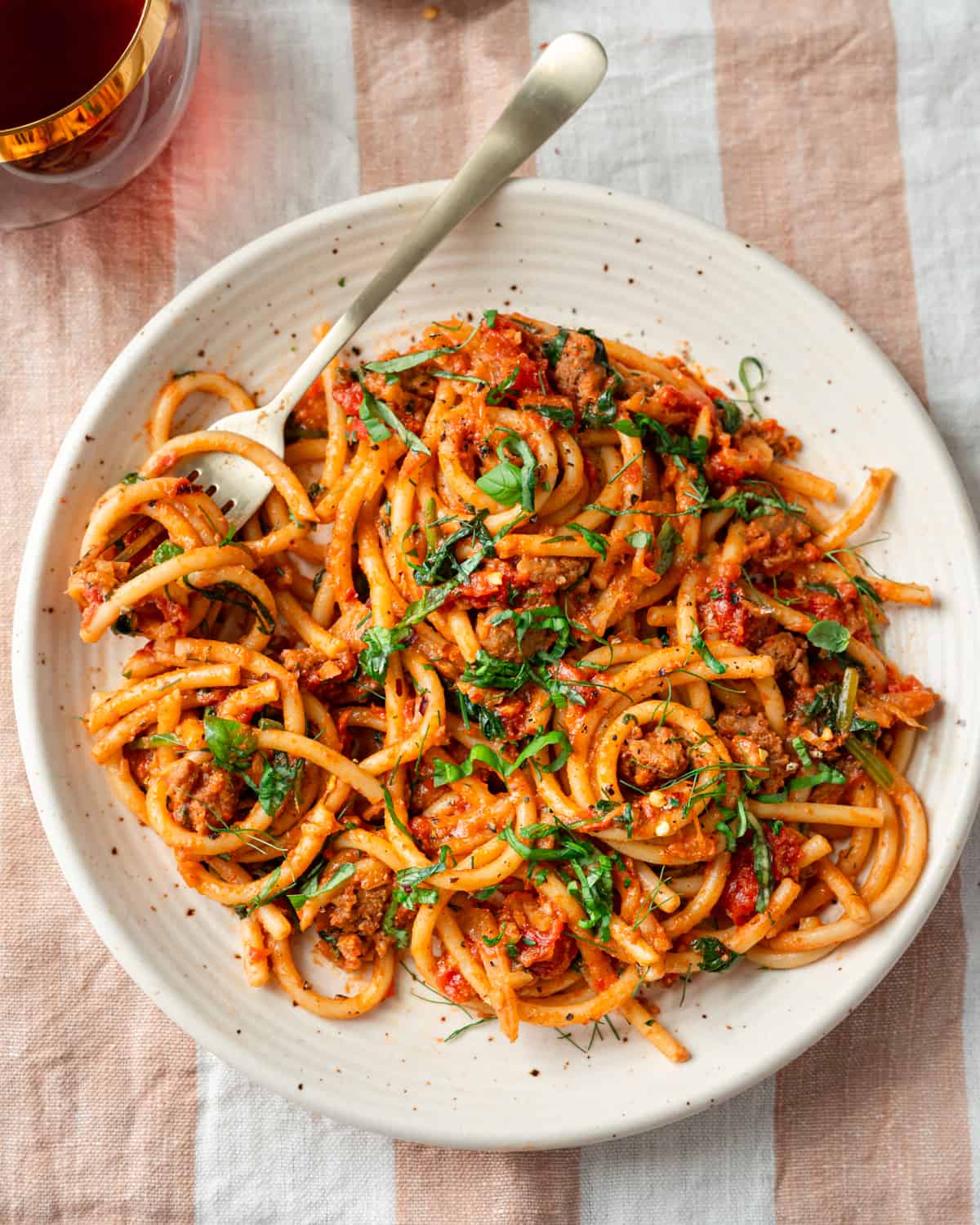 bucatini pasta with sausage tomato sauce, garnished with basil on a speckled plate on a striped napkin.