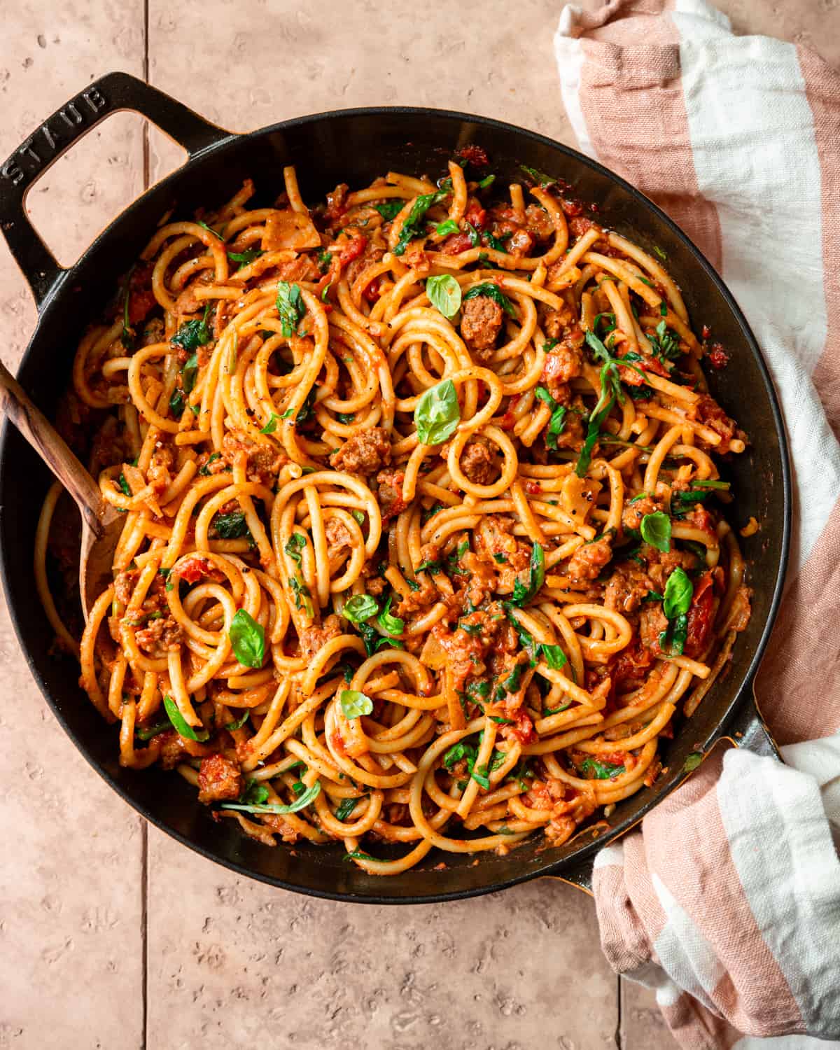 bucatini pasta with sausage tomato sauce, garnished with basil in a cast iron skillet on a tiled surface.