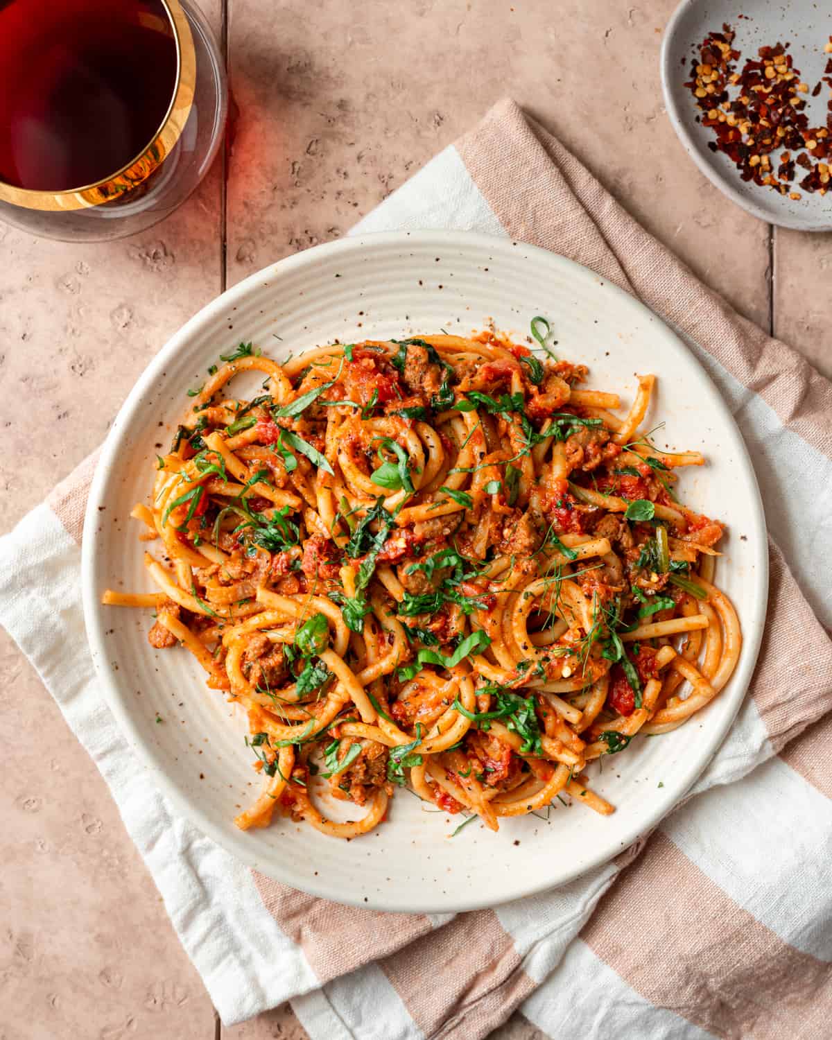 bucatini pasta with sausage tomato sauce, garnished with basil on a speckled plate on a striped napkin.