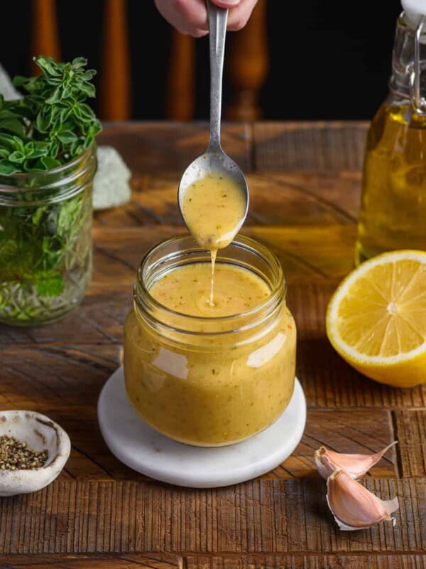 person taking a spoon from a glass jar of vinaigrette on a wooden table.