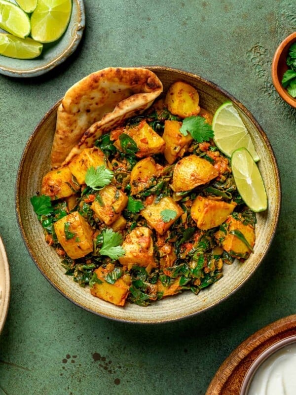 bowl of saag aloo with naan and lime wedges on a dark green table.