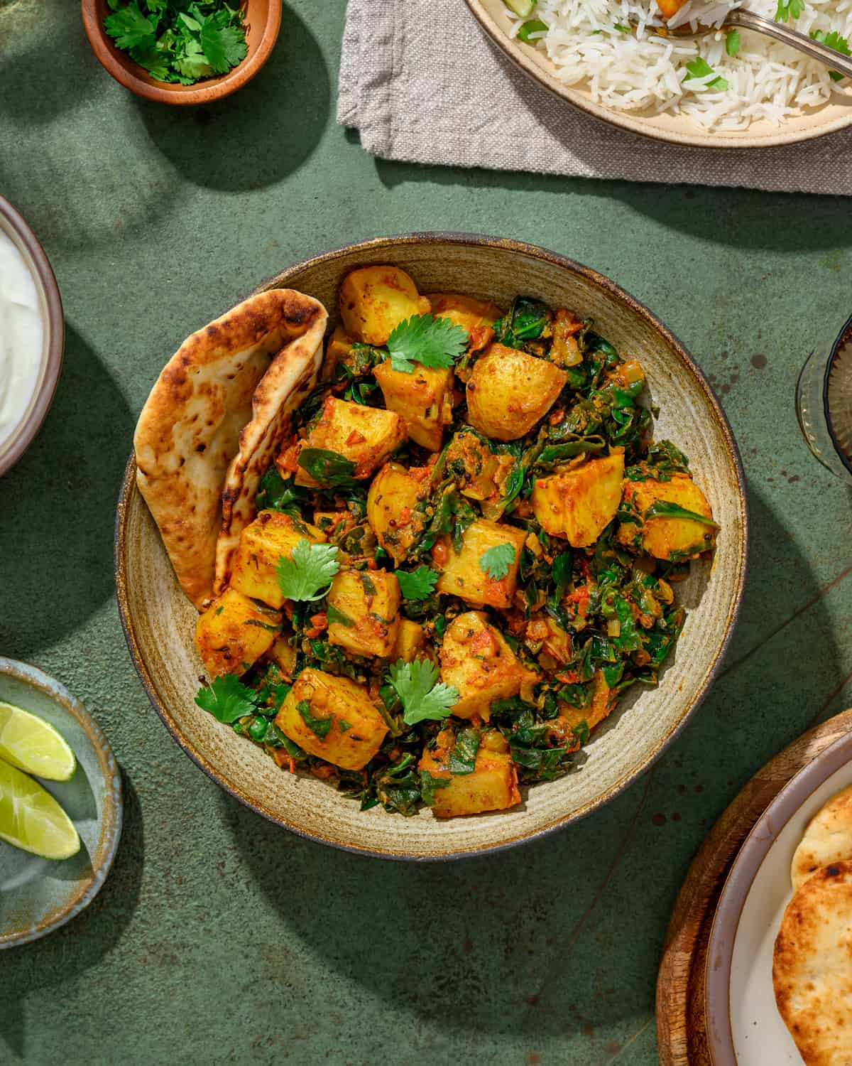 bowl of saag aloo with naan and lime wedges on a table.