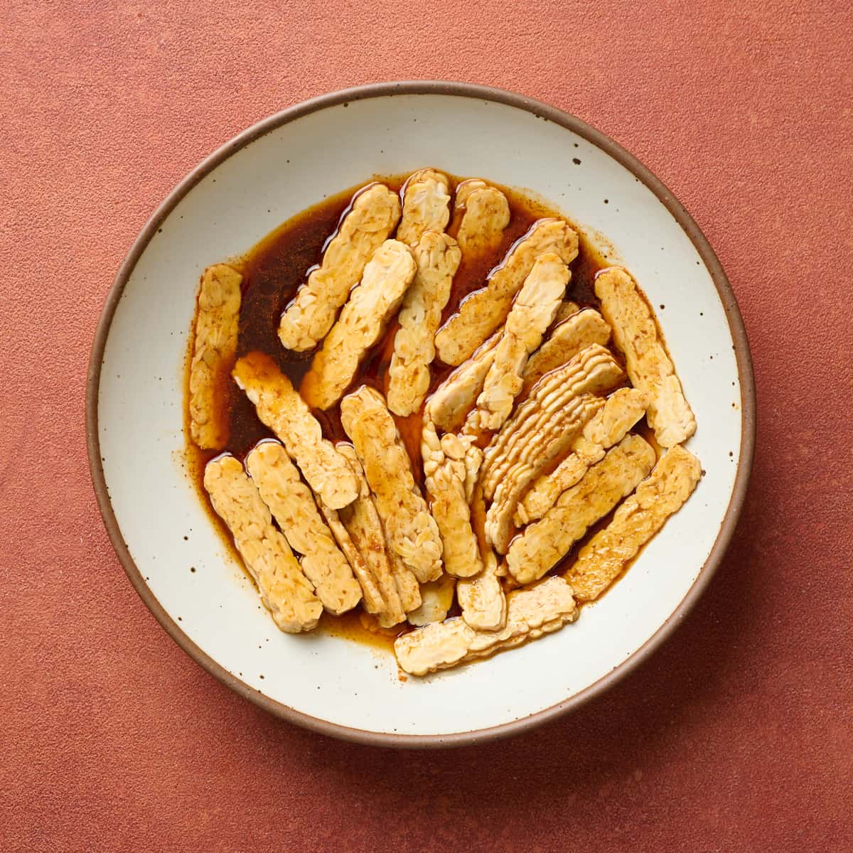 Tempeh strips and marinade mixed in a large white bowl.