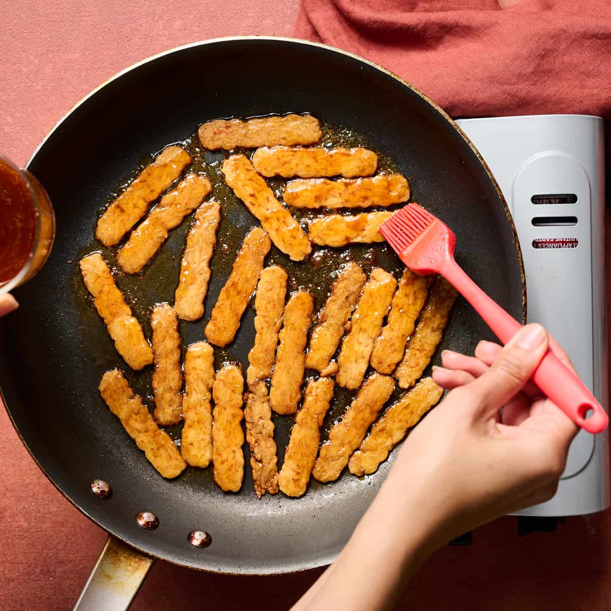 Woman's hand brushing reserved marinade tempeh slices cooking in a frying pan.