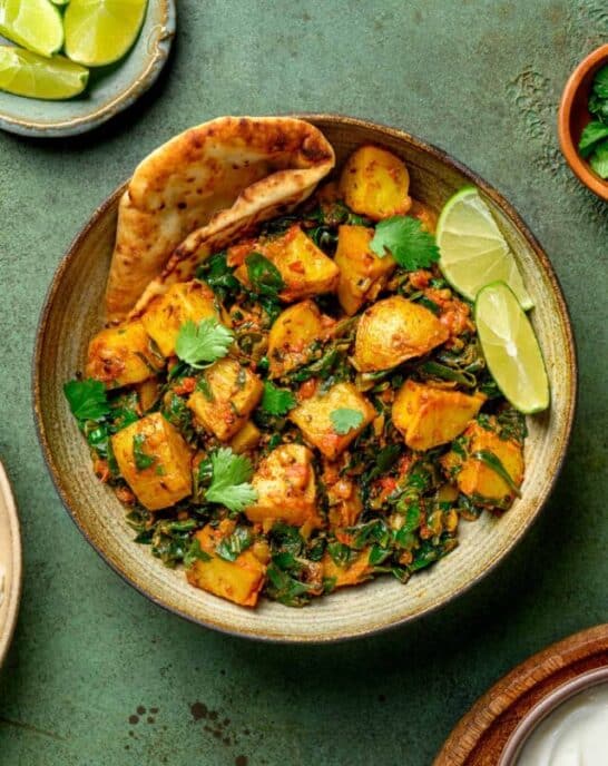 bowl of saag aloo with naan and lime wedges on a dark green table.
