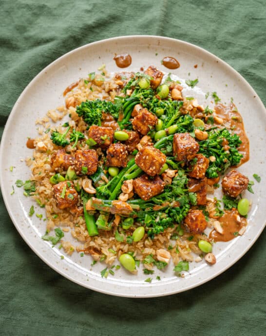 baked peanut tofu with broccolini and edamame on top of brown rice on a ceramic plate.