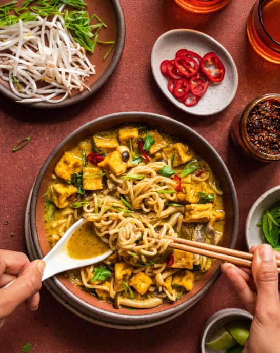 Malaysian curry soup with women's hands digging in with a spoon and chopsticks.