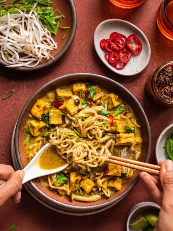 Malaysian curry soup with women's hands digging in with a spoon and chopsticks.