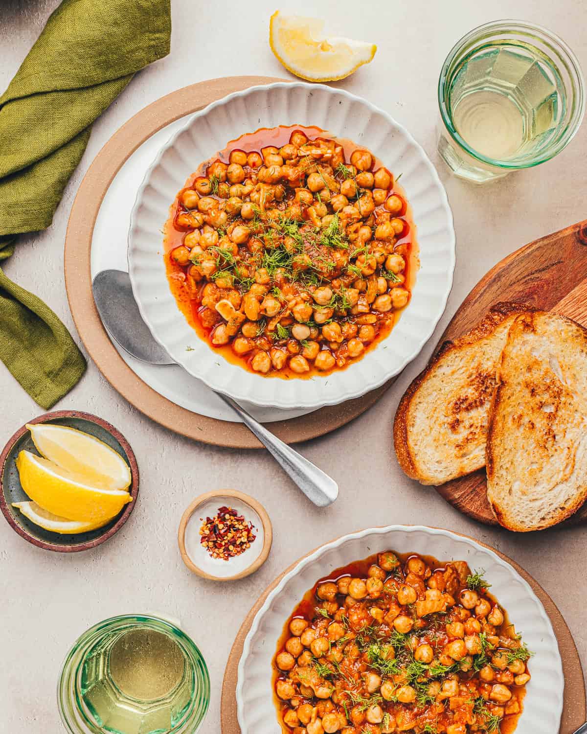 Two bowls of braised chickpeas on a table next to bread, glasses of water and sliced lemons.