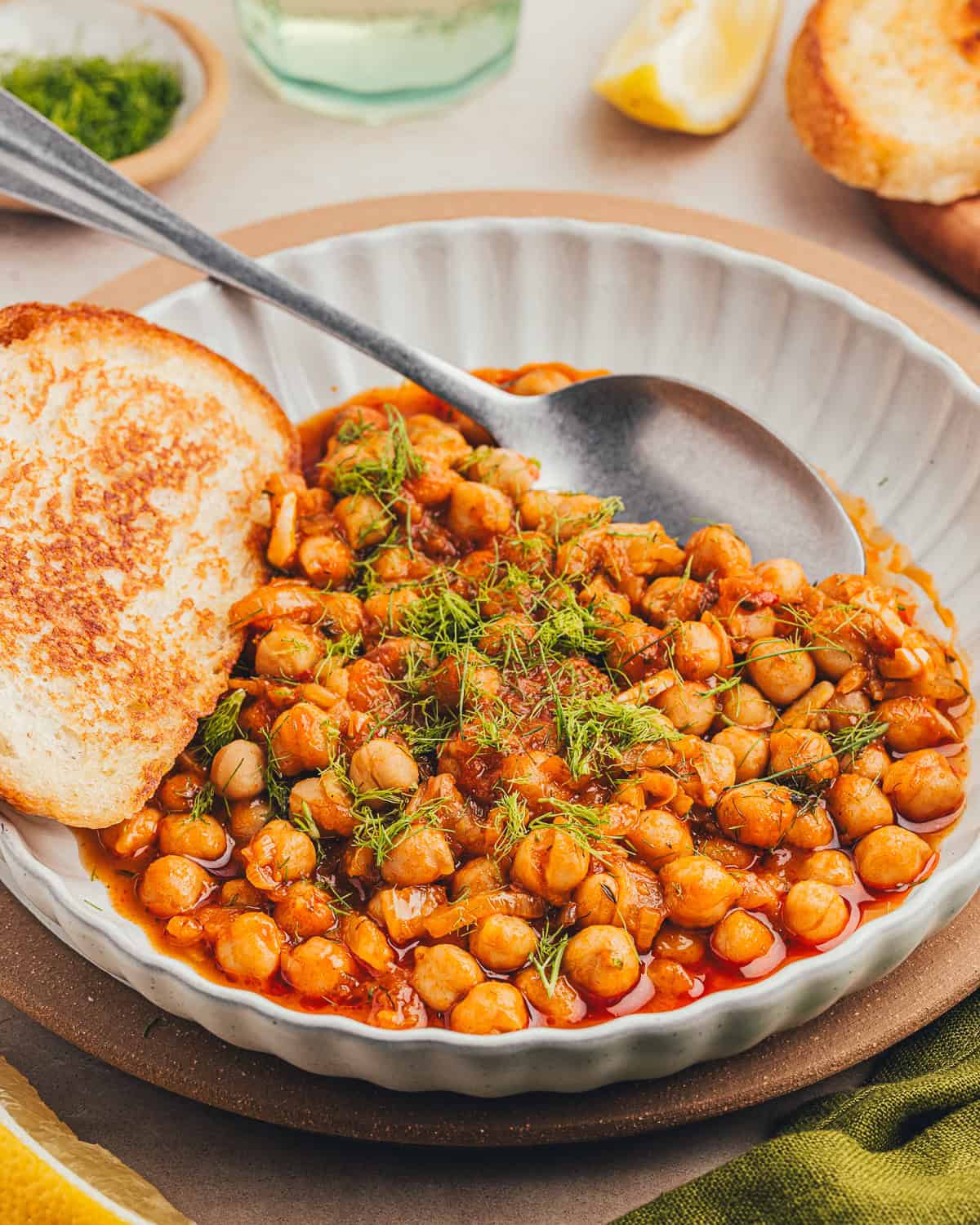 Close up shot of bowl of braised chickpeas with a slice of bread and a spoon.