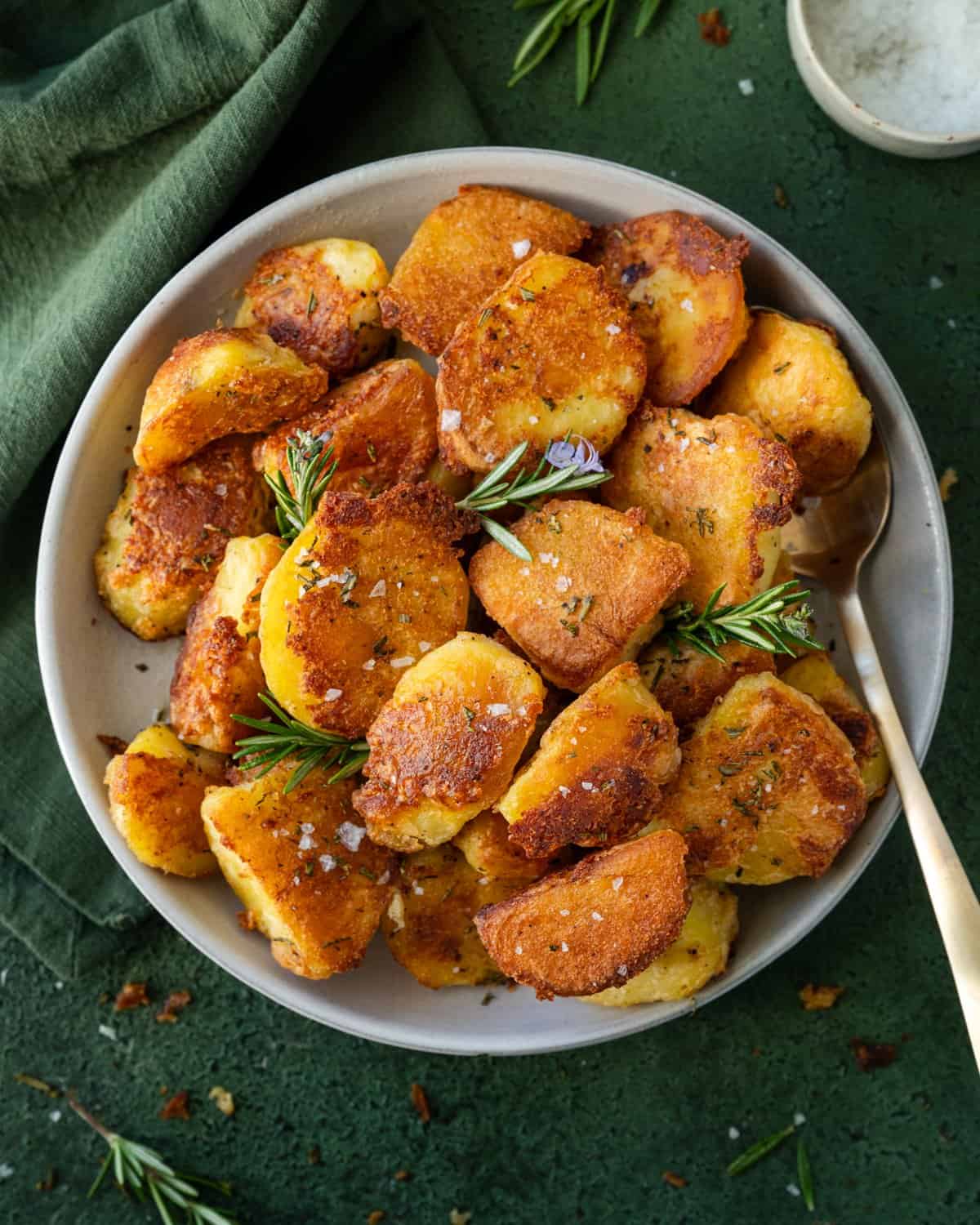 Crispy roasted potatoes with small rosemary sprigs and serving spoon on a tablecloth.