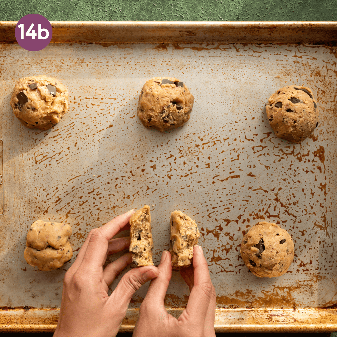 woman's hands holding a cookie dough ball torn in half on a sheet pan.