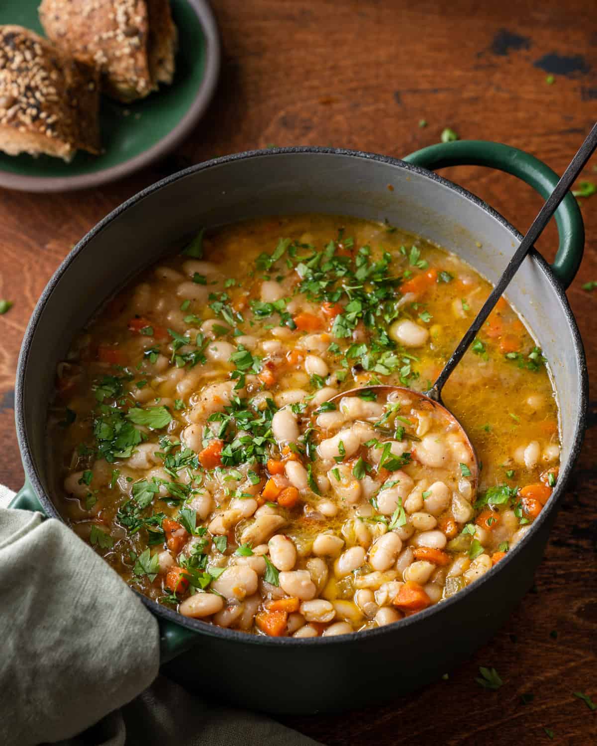 ladle digging into a green dutch oven with white bean soup on wooden table.