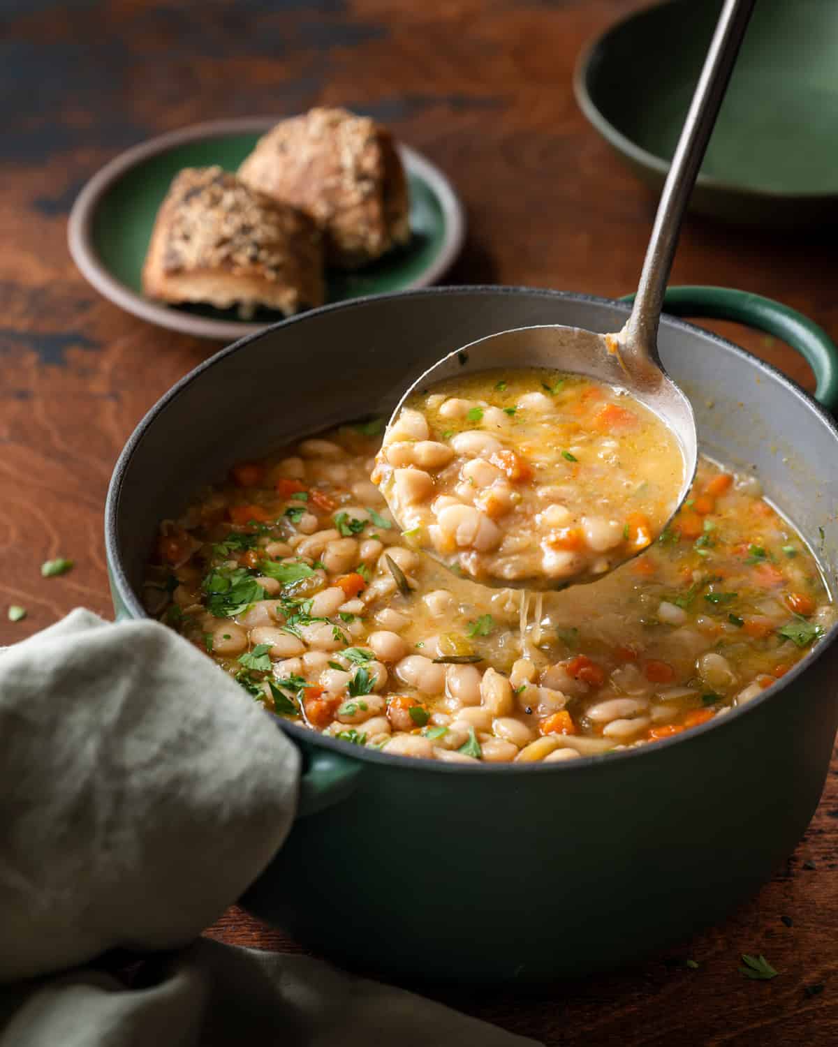 a ladle scooping up white bean soup from a green dutch oven on a wooden table. 