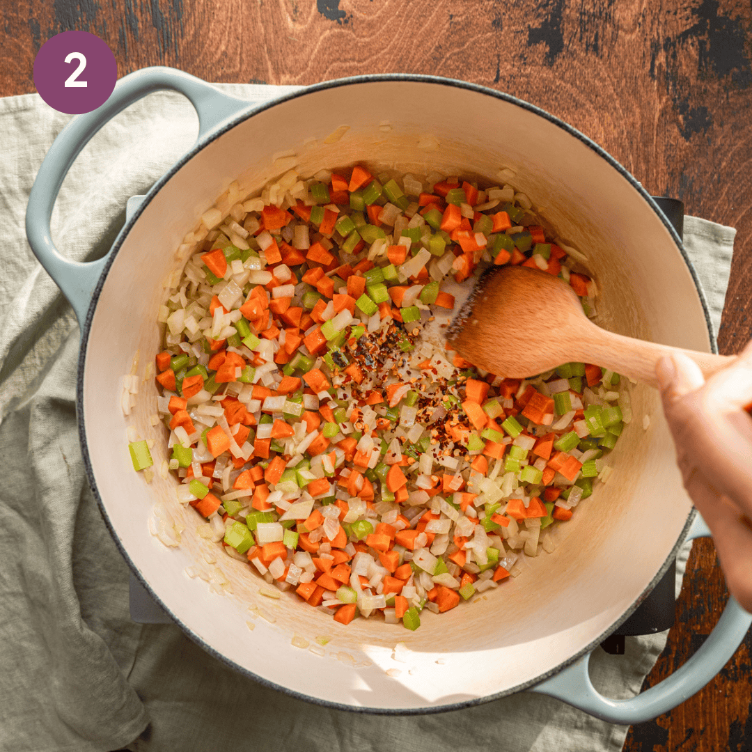 wooden spatula stirring diced carrots, onions, and celery with red pepper flakes in a dutch oven. 