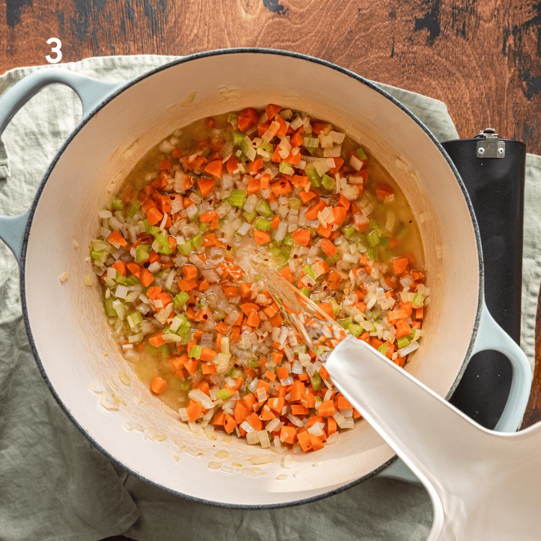 pouring water into a dutch oven with onions, carrots, and celery sauteing.