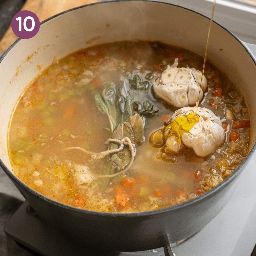 drizzling olive oil on top of whole garlic heads sitting on top of a soup in a dutch oven.