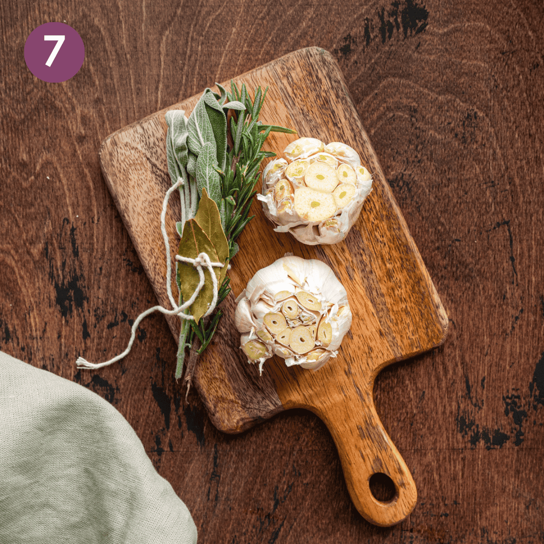 two heads of garlic with tops sliced off and a bouquet garni on a small wooden cutting board. 