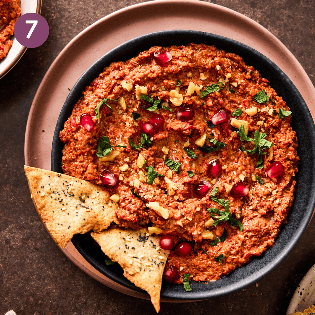 Muhammara in a shallow serving bowl with herbs, pomegranate seeds, walnut chunks and pita chips.