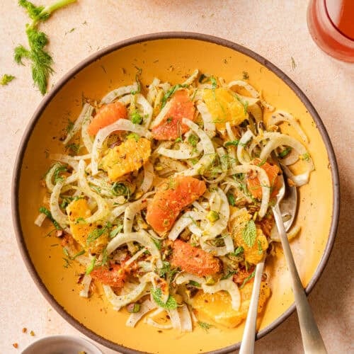 fennel orange salad in a yellow bowl with gold silverware on a pink surface.