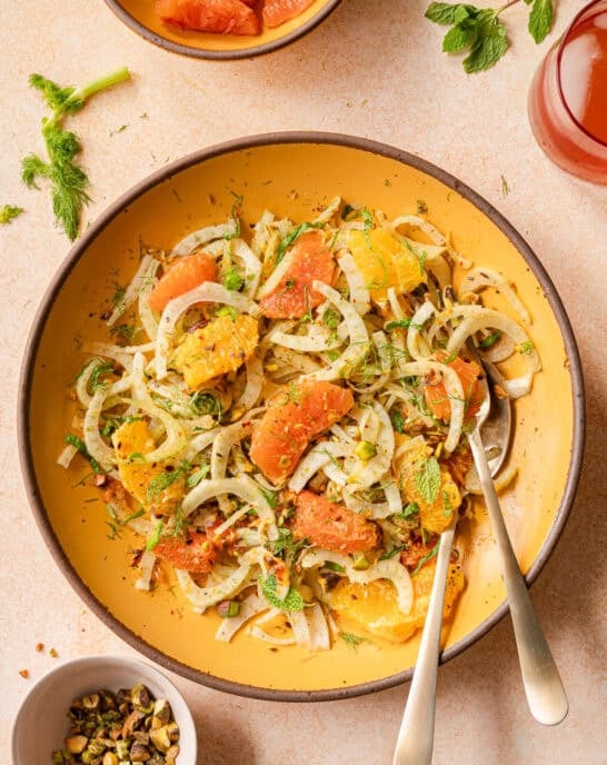 fennel orange salad in a yellow bowl with gold silverware on a pink surface.