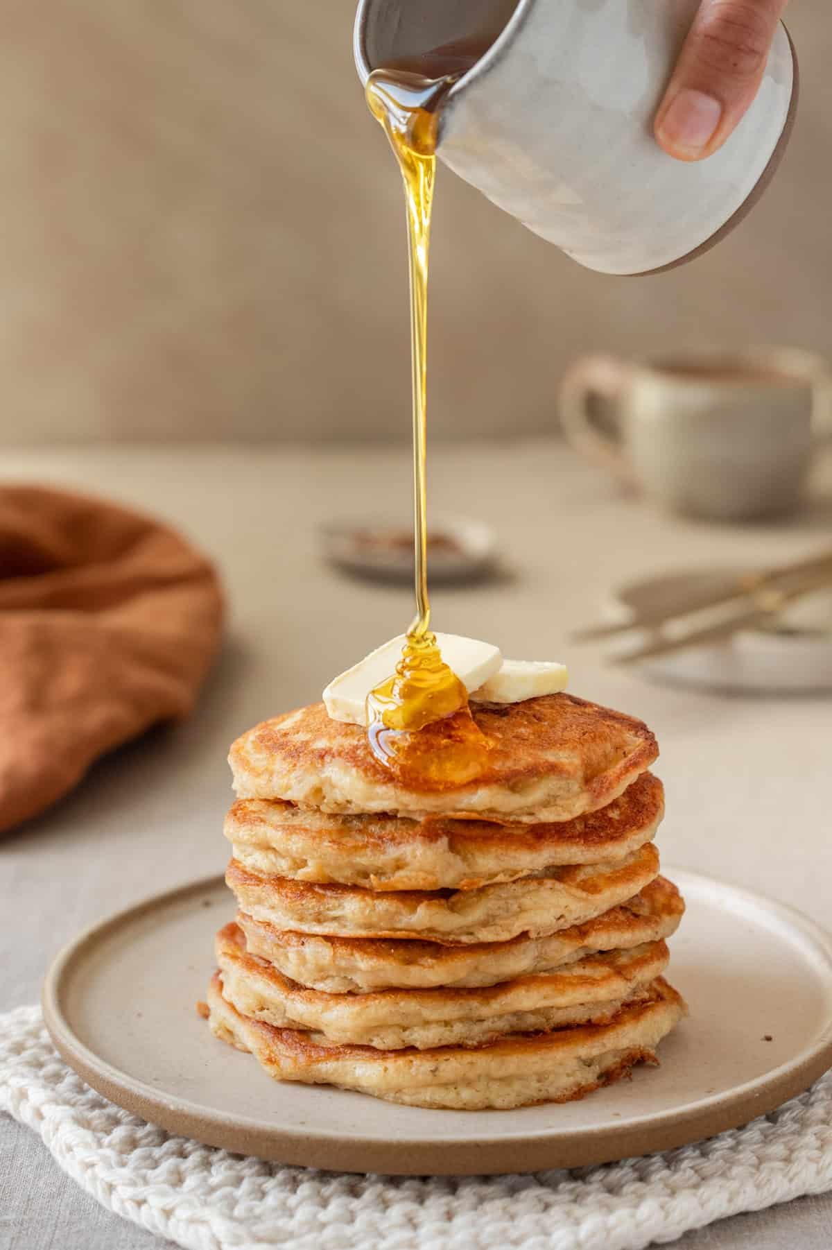 Side view of person pouring maple syrup onto a tall stack of banana pancakes on a plate.