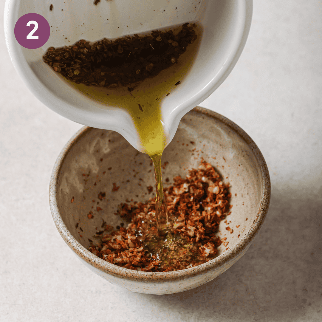person pouring hot oil over garlic and chili flakes in a small bowl.