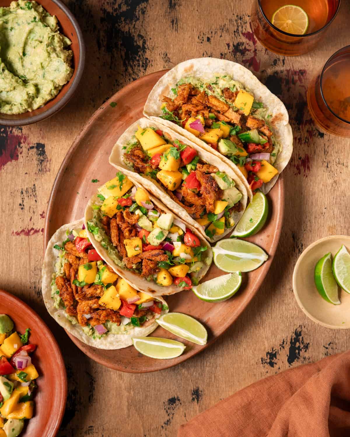 a table with four vegan tacos on a large plate next to ingredient bowls.