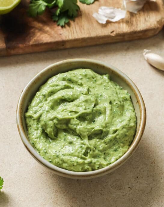 bowl of avocado crema on a table next to a cutting board with ingredients.