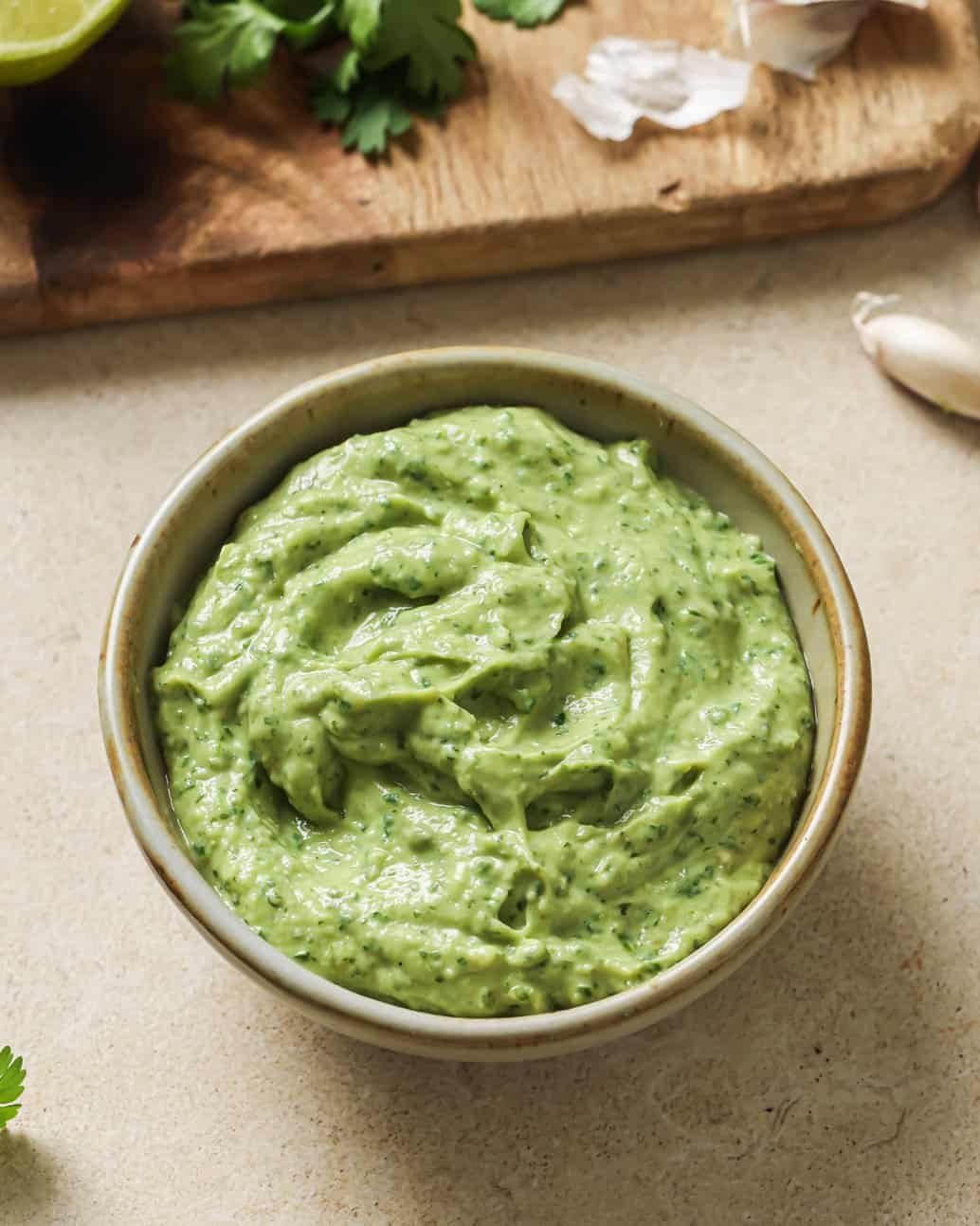 bowl of avocado crema on a table next to a cutting board with ingredients.