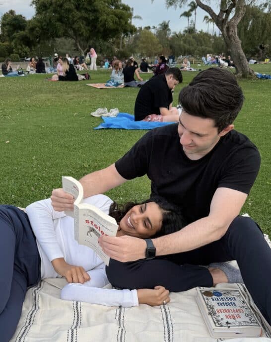 Max holding up a book for Nisha as she lies down on a blanket in the park.