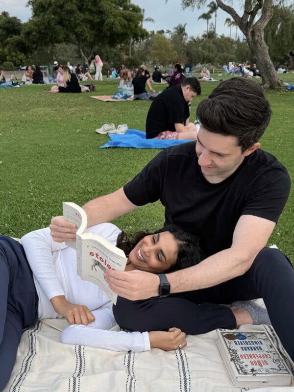 Max holding up a book for Nisha as she lies down on a blanket in the park.