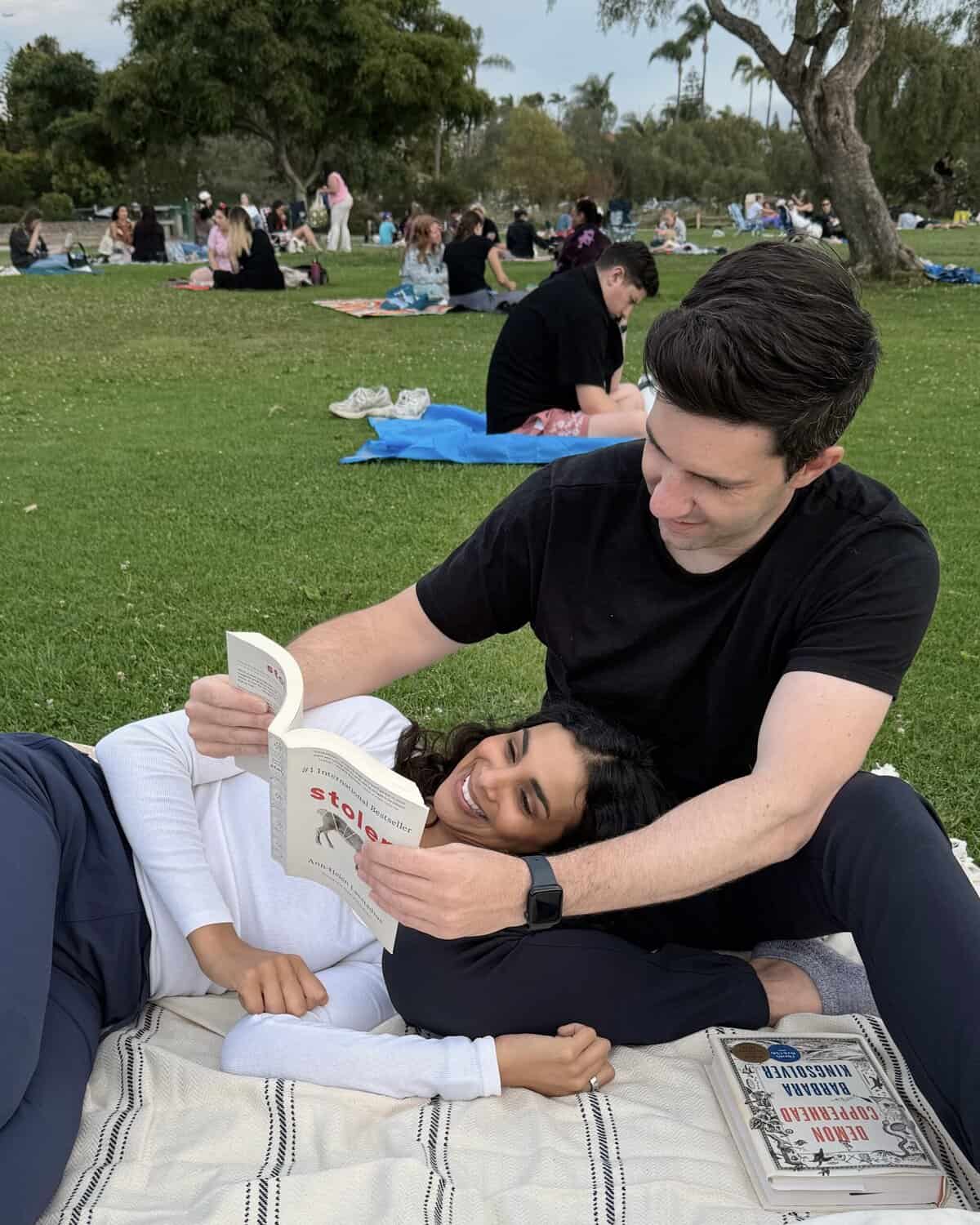 Max holding up a book for Nisha as she lies down on a blanket in the park.