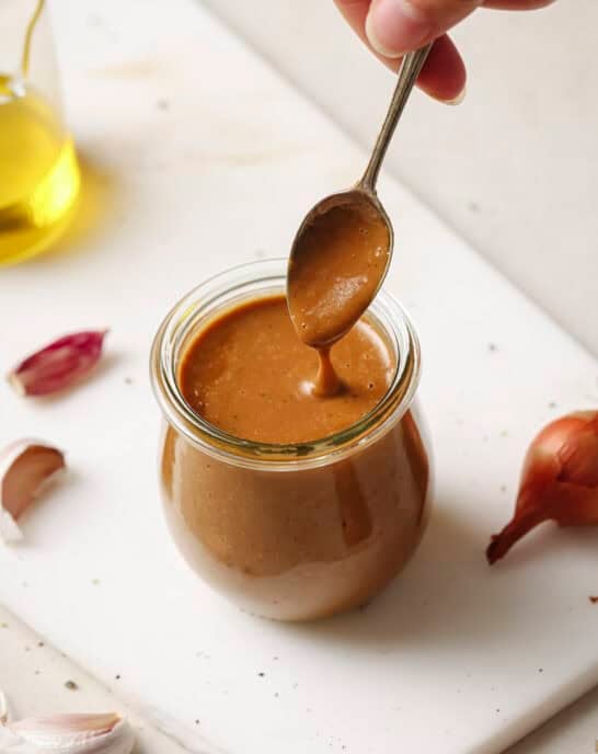 Woman's hand spooning creamy balsamic vinaigrette from a jar on a white surface.