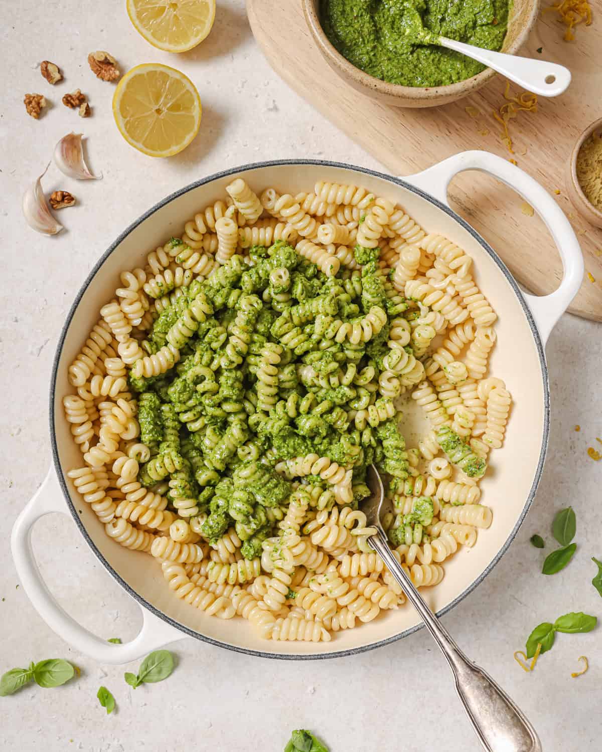 large pot of pasta with spinach pesto surrounded by various ingredients used in pesto on a table.