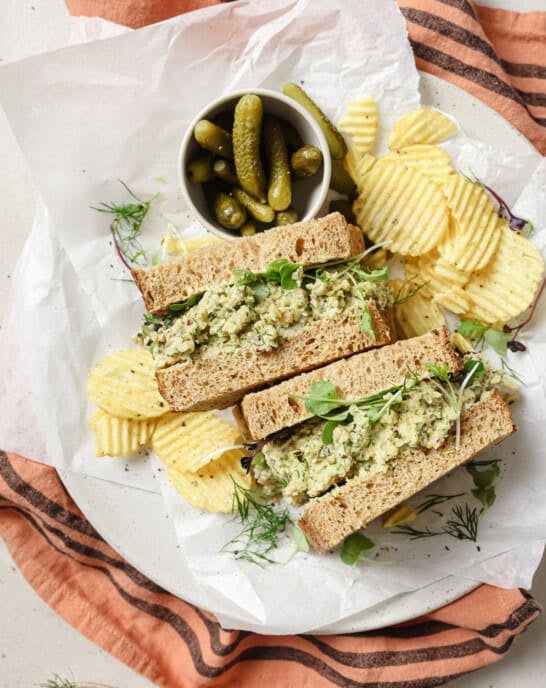 two chickpea salad sandwich halves on a plate with potato chips and cornichons.