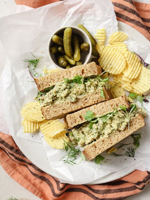 two chickpea salad sandwich halves on a plate with potato chips and cornichons.