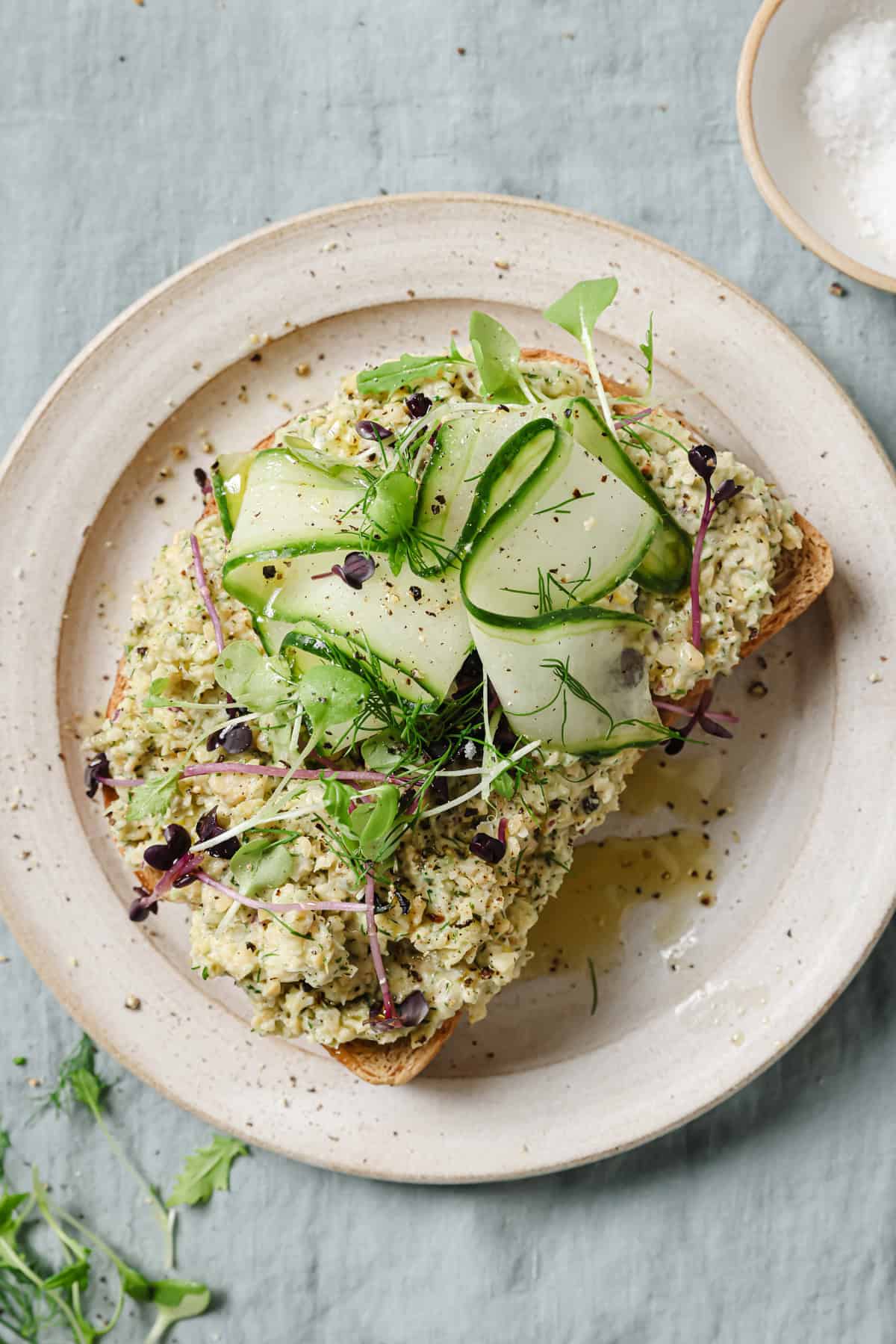 open faced chickpea salad sandwich topped with cucumber ribbons and fresh herbs on a plate.