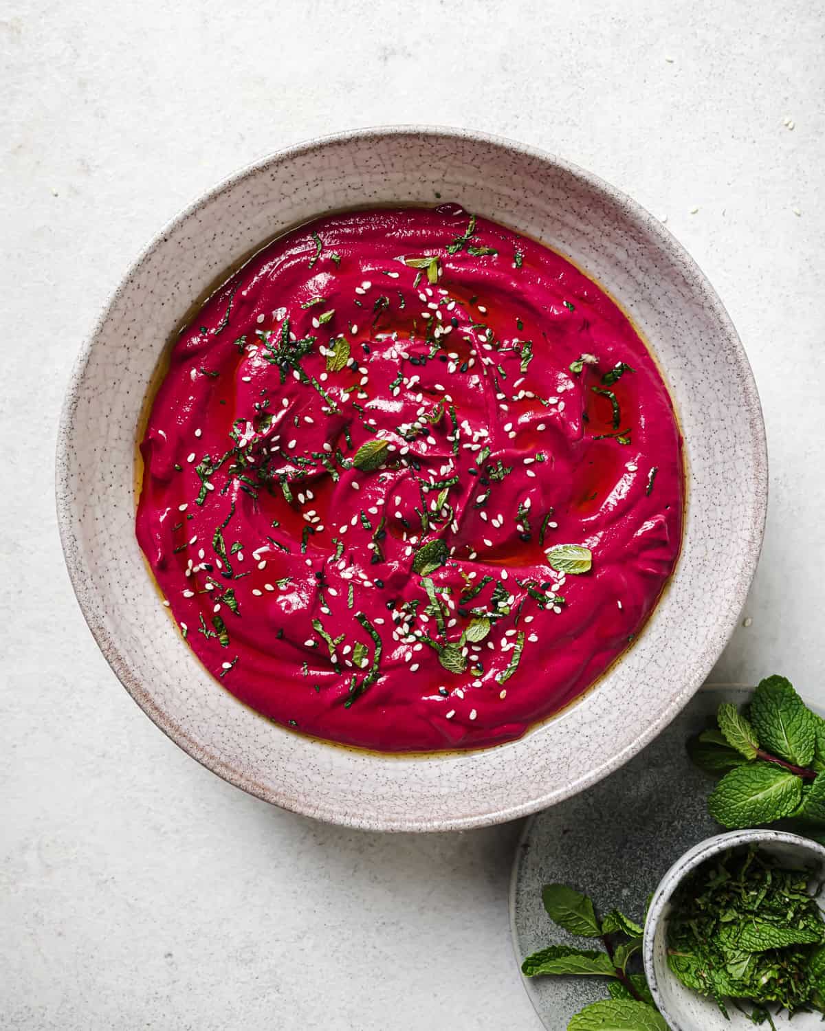 Overhead shot of beet dip with sesame seeds and mint next to a small plate of chopped mint.