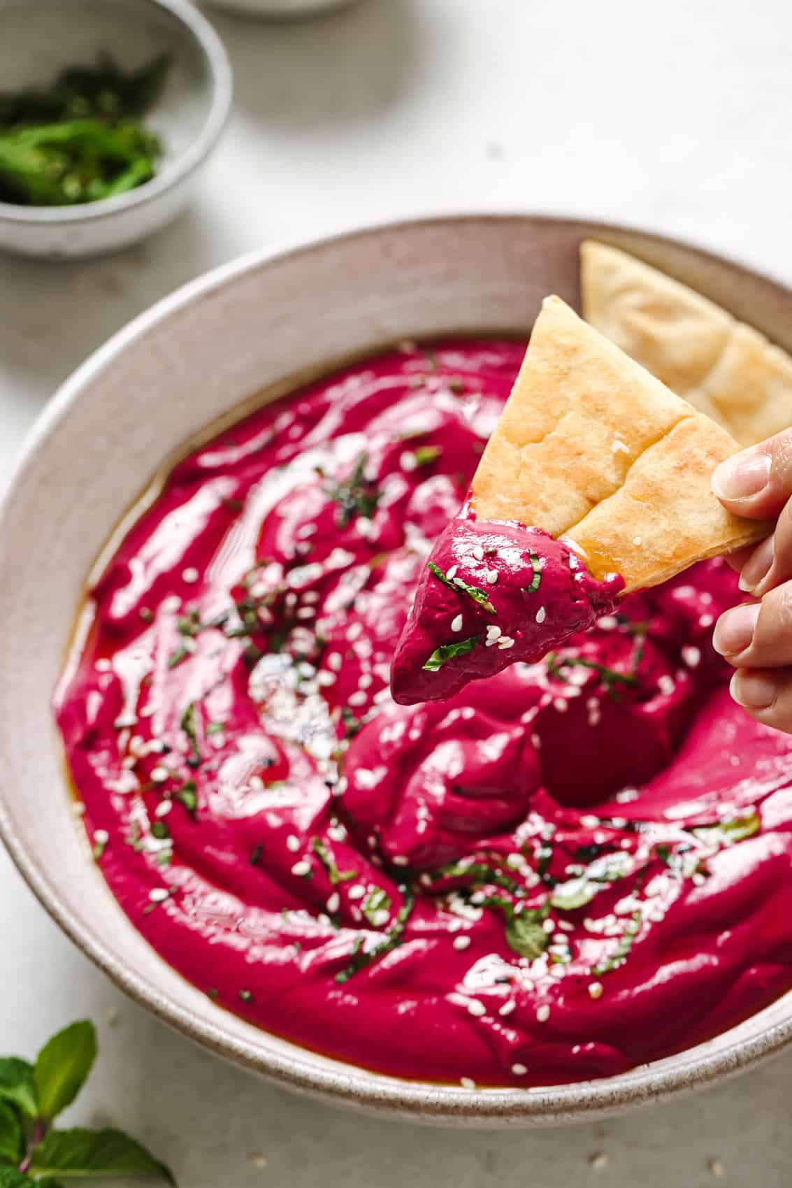 a person holding a triangle of pita bread with beet dip above a bowl of the dip.