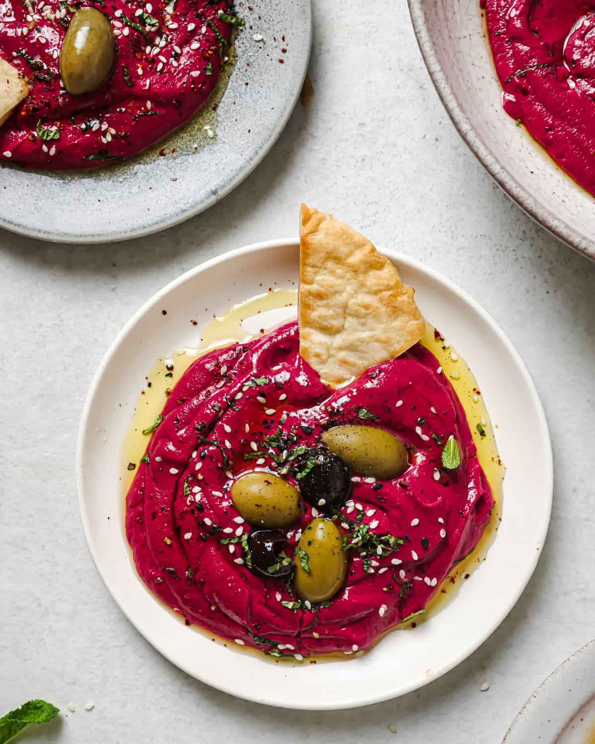 small plate of beet dip topped with olive oil, olives, sesame seeds and mint with a pita triangle.